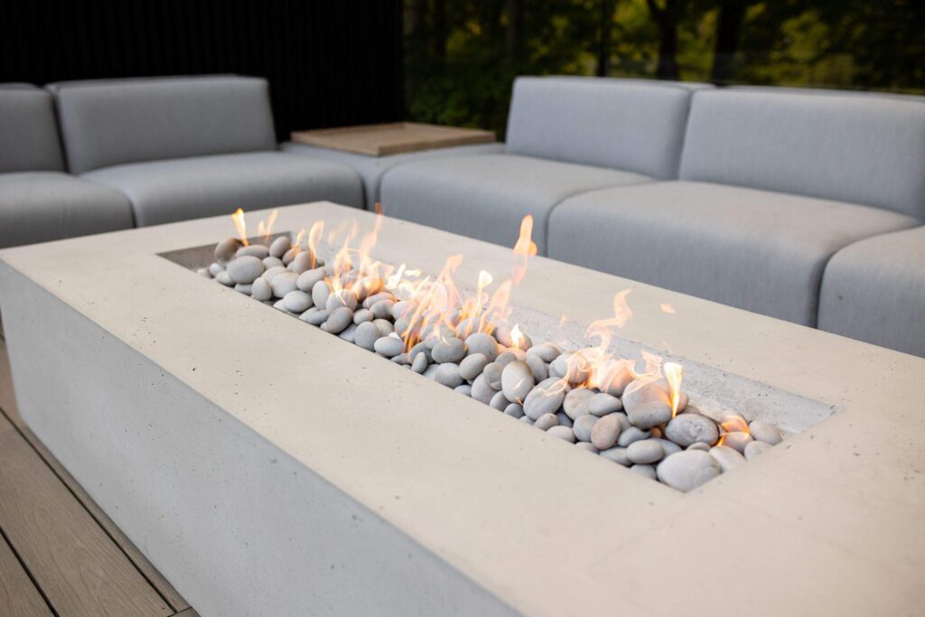 Modern outdoor natural gas fire pit with a rectangular concrete base and smooth stone accents, surrounded by minimalist grey seating, offering a stylish and inviting atmosphere for outdoor gatherings.