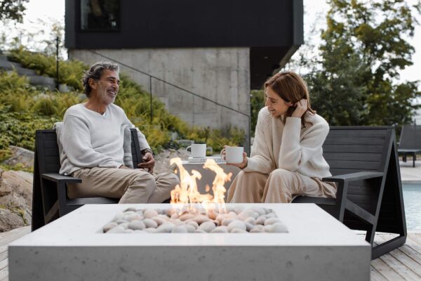 Two people enjoying a cozy moment with hot beverages by a modern concrete outdoor fire pit, surrounded by lush greenery and a contemporary house in the background.