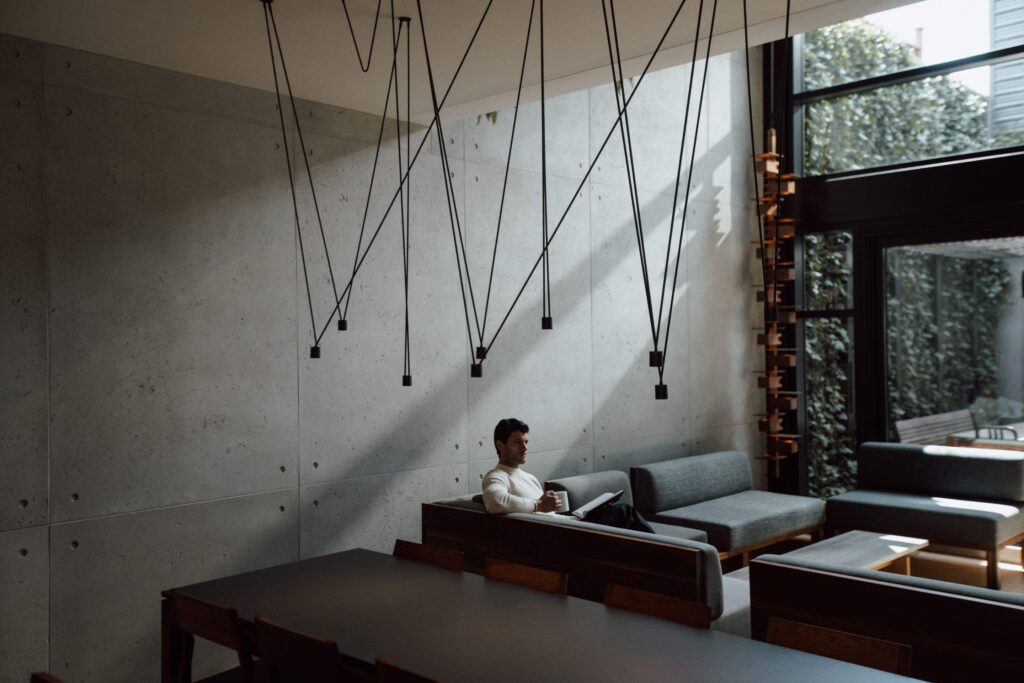 A stylish living room featuring a man sitting on a gray sectional sofa with a book. The space has high concrete walls, large windows with an outdoor view, and unique pendant lights with black wires forming geometric shapes hanging from the ceiling. The room exudes a cozy yet modern ambiance.