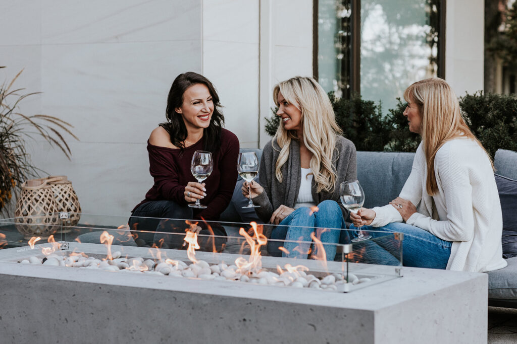 Three middle-aged women sitting together drinking wine. Outdoor patio space with outdoor furniture and a concrete fire pit. 